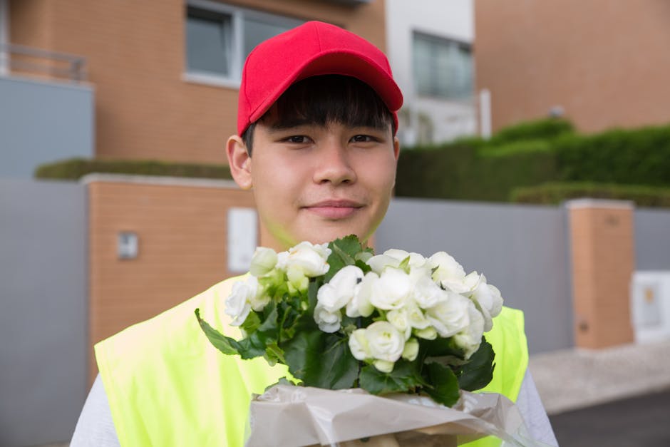 A young male flower delivery courier, dressed in a neon yellow vest and red cap, holding a bouquet of fresh white flowers with green leaves in front of him. The bouquet is wrapped in white paper, show