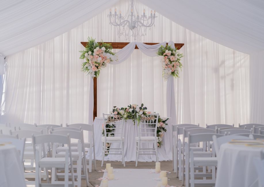A wedding ceremony setup inside a white marquee features a wooden arch decorated with large, soft pink and white floral arrangements including roses and peonies, complemented by green foliage. The bac