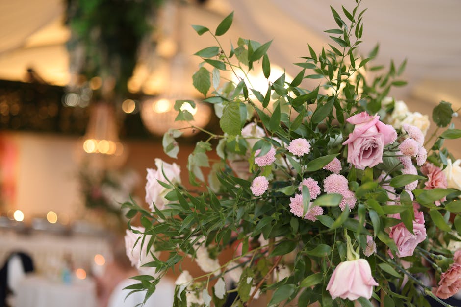 A floral arrangement featuring soft pink roses with full, layered petals and delicate pink pom-pom-like blooms, interspersed with lush green foliage of various elongated and pointed leaves. The flower